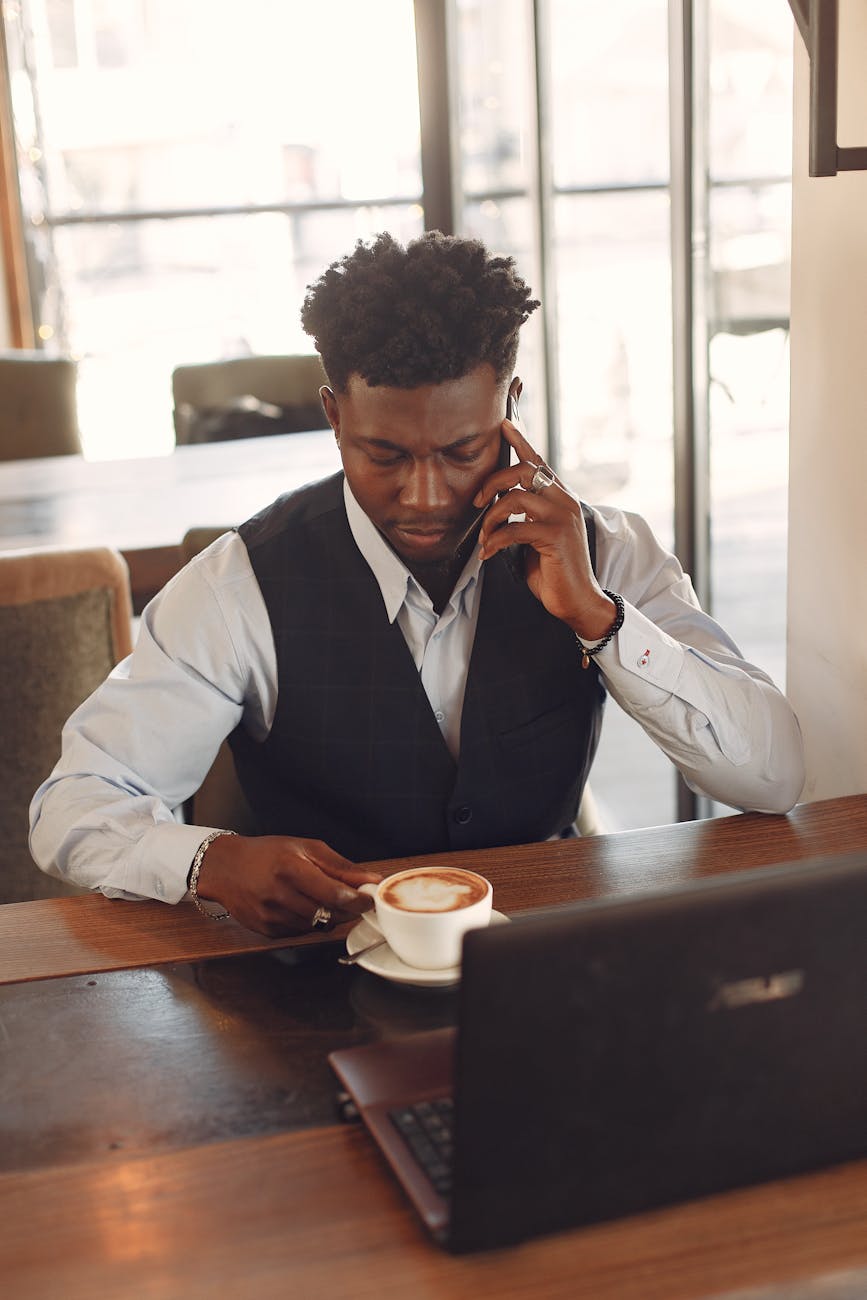 serious african american businessman talking on smartphone during coffee break using laptop in cafe